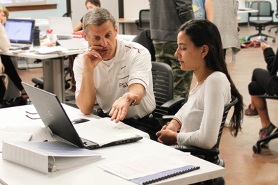 A professor seated at a desk gestures to a computer during a conversation with a student.