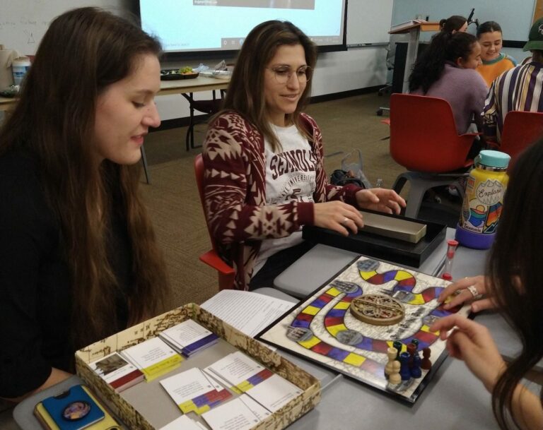Students sit at a table. They are playing a colorful board game.