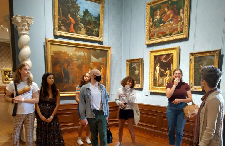 College students and their professor stand in a museum gallery room with blue walls covered in paintings.