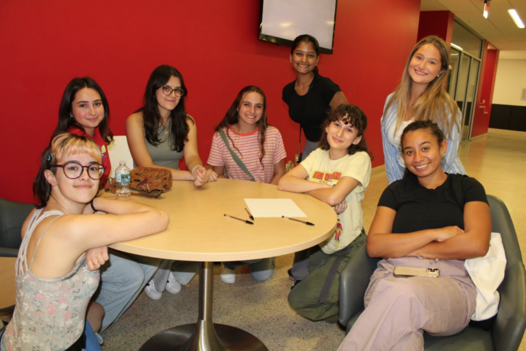 A group of college students sit at a table in front of a bright red wall. They are posing for a photo.