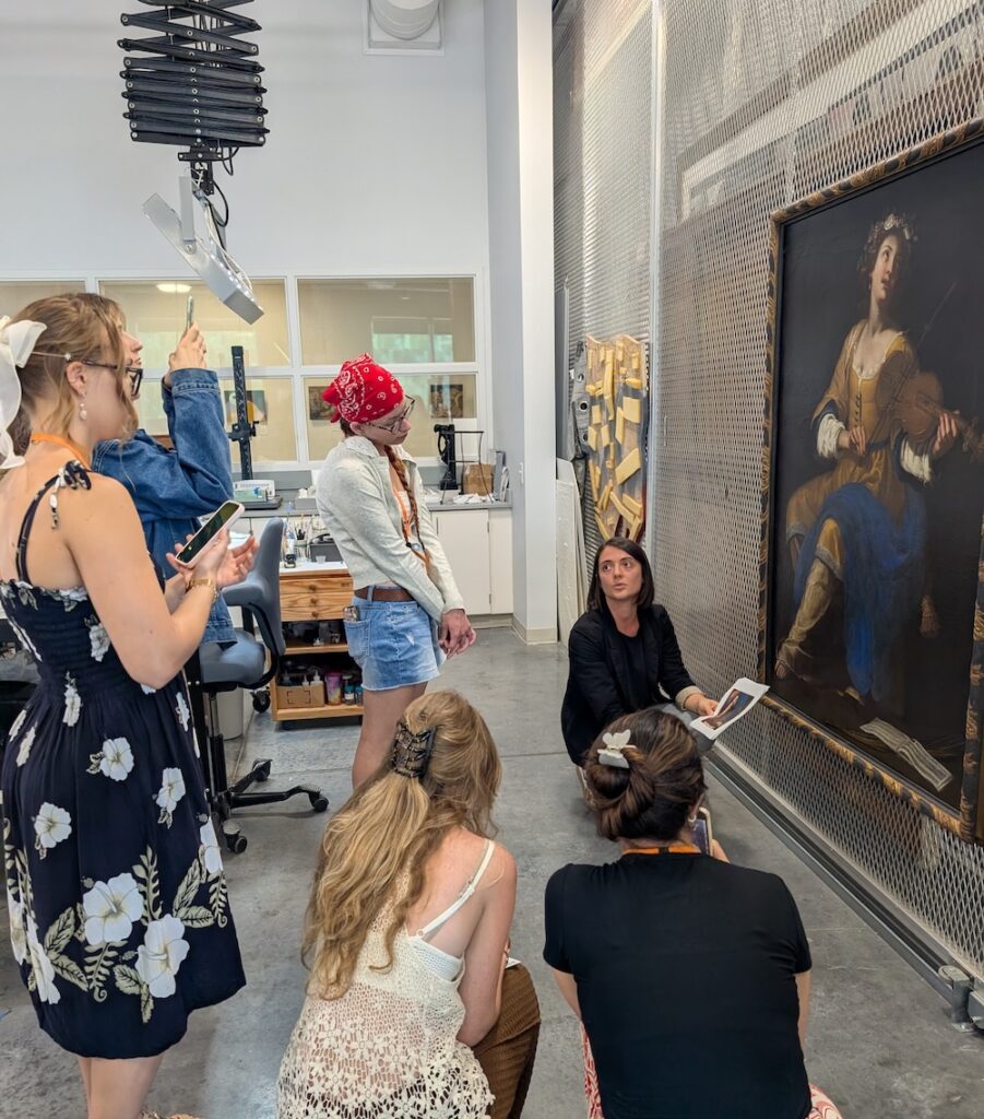 A woman, crouched down in front of a large oil painting, speaks to a crowd of engaged students.