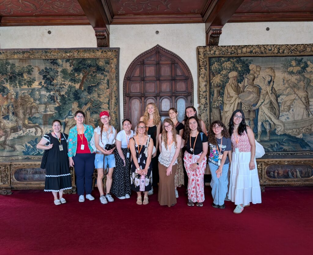 A group poses for a photo in front of large historic doors and tapestries